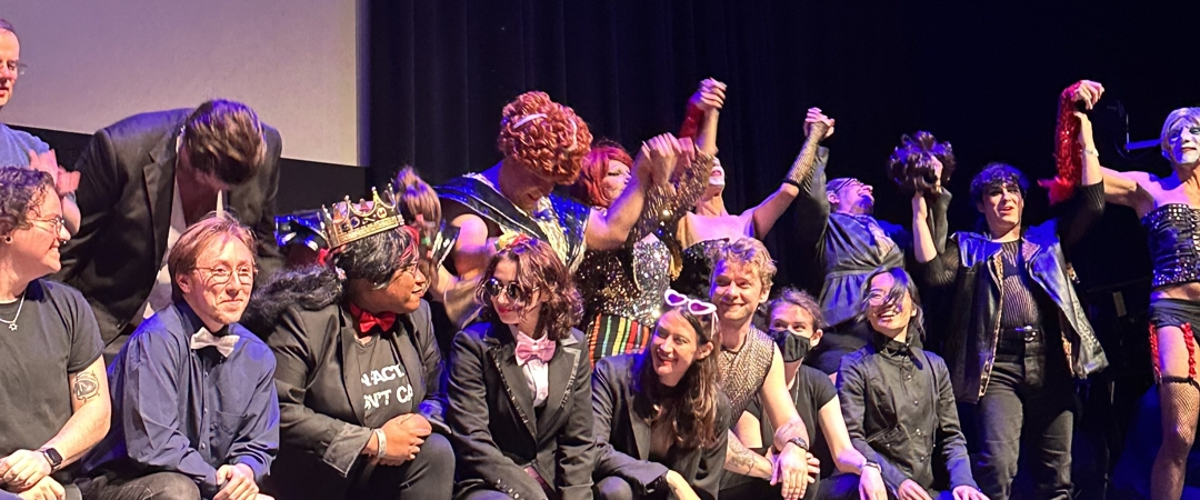 Shadowcast actors and crew taking a cast photo on stage at the end of a performance. The Rocky Horror Picture Show is playing on the screen behind them.